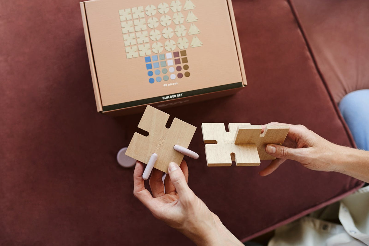 Adult hands joining two BAVVIC beech-wood pieces with silicone connectors next to a BAVVIC set box on a sofa